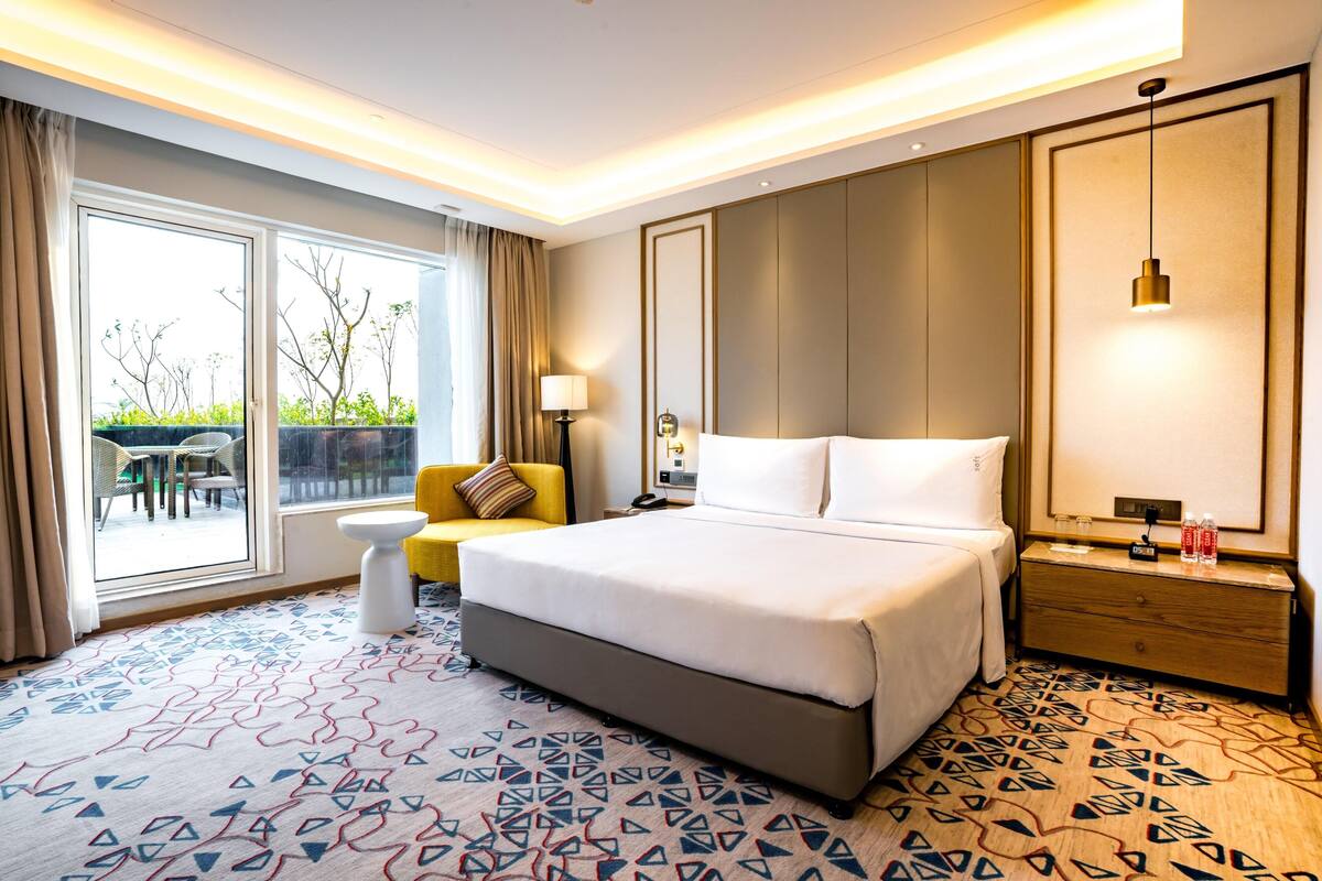 A hotel room with a large bed, a bedside table with a lamp, a small sofa, and a view of the outdoors.