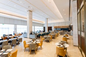 A spacious hotel dining area with multiple tables and chairs, a kitchen in the background, and a large window providing natural light. - Holiday Inn Lucknow Airport (Lucknow)