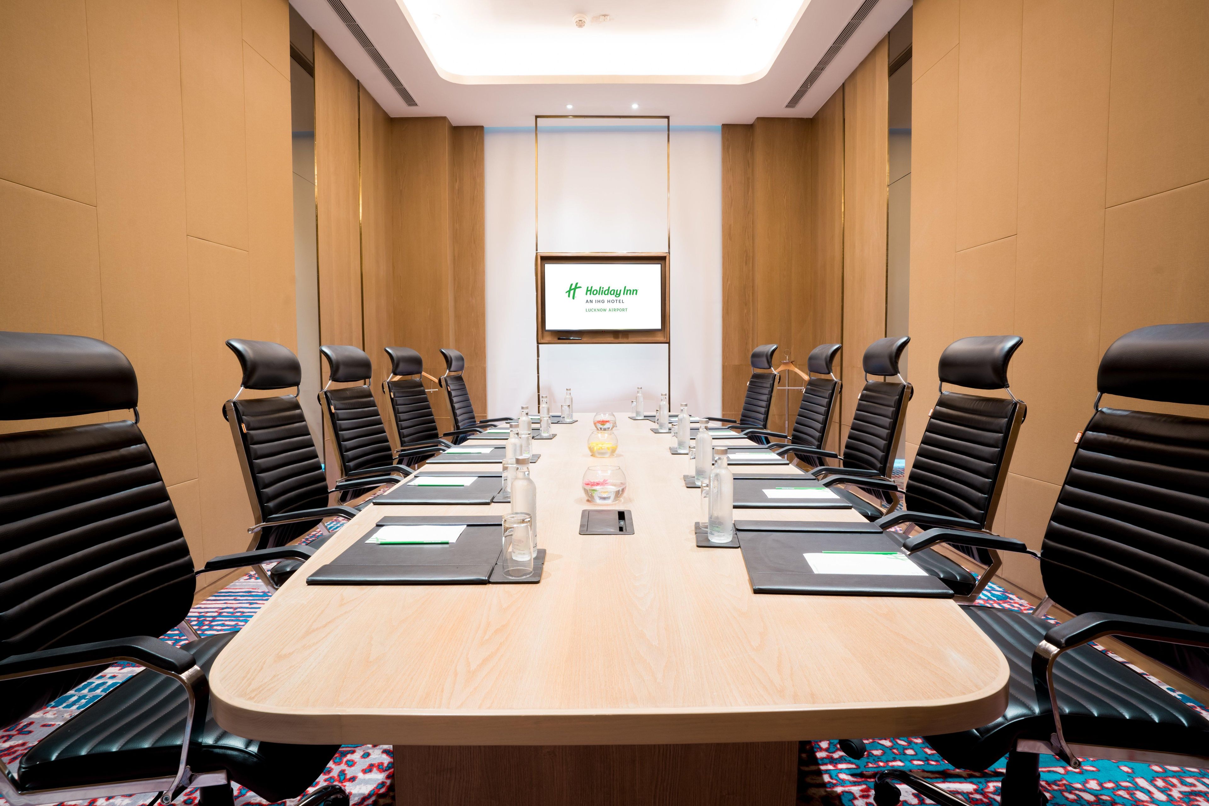 Een vergaderzaal met een lange tafel, zwarte stoelen en een scherm waarop het Holiday Inn logo staat.