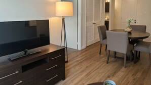 A modern living room with a flat-screen TV, dark wooden entertainment unit, a floor lamp, and a dining area with a round table and chairs.