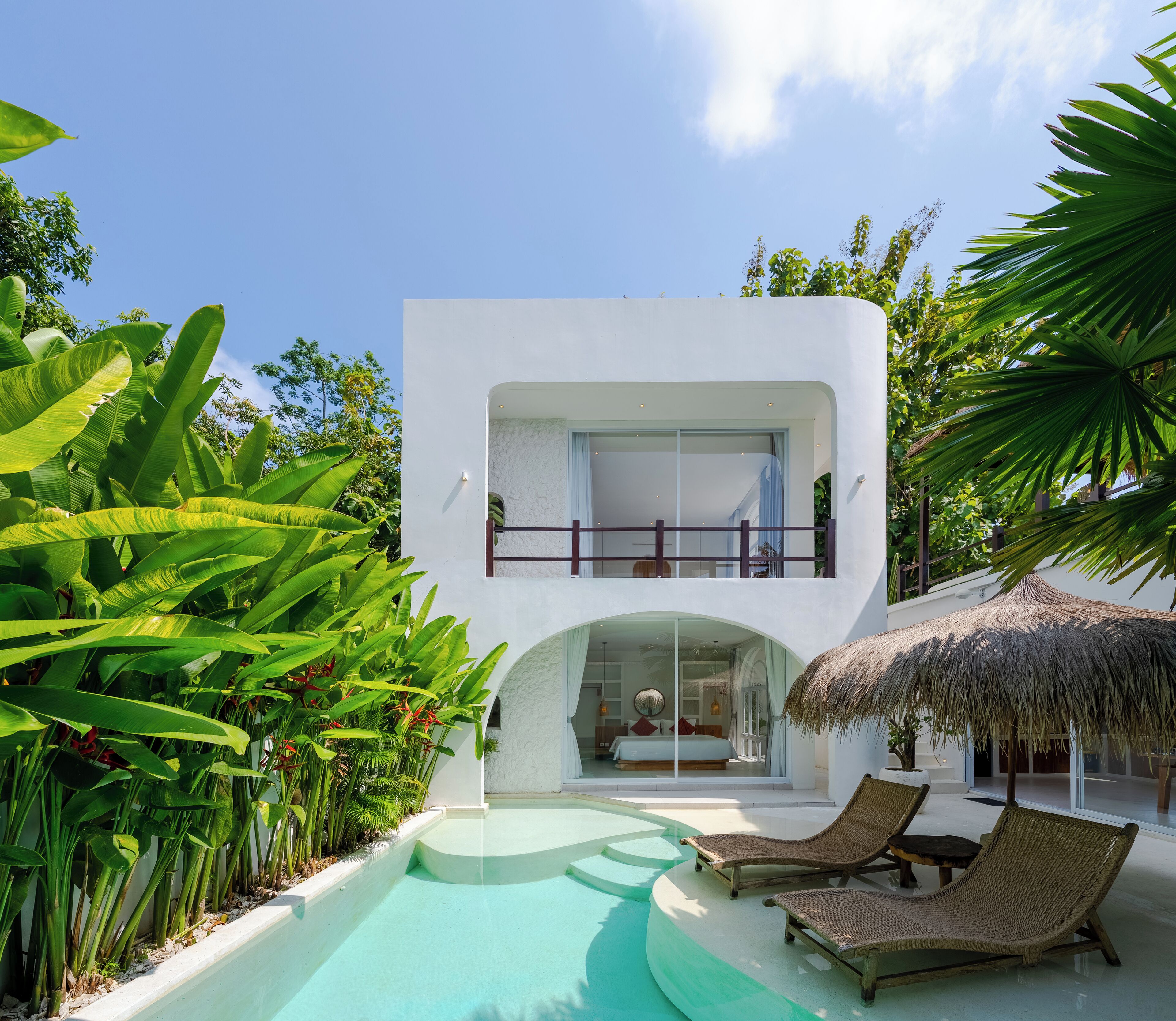 Un edificio moderno de dos pisos con balcón de vidrio, piscina y plantas tropicales.