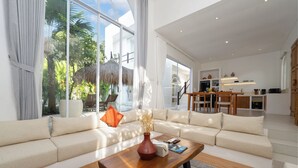 Un soggiorno moderno con divano, tavolino e vista su un giardino tropicale attraverso ampie finestre.