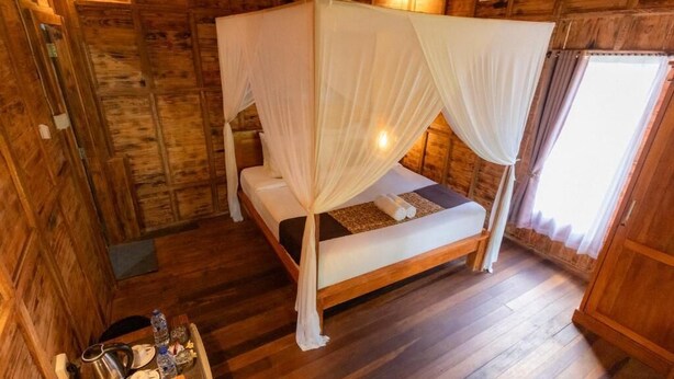 A wooden cabin room with a bed under a canopy, a small table with a kettle and plates, and a window with curtains.