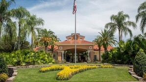 A building with a red-tiled roof, an American flag, and a well-maintained garden with palm trees and yellow flowers. - 4bd Villa Near Epic Universe And Disney With Private Pool 4 Bedroom Townhouse (Kissimmee)