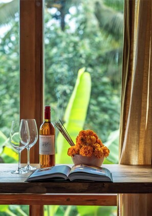 Uma mesa de madeira com uma garrafa de vinho, duas taças, uma tigela de flores de calêndula e um livro aberto perto de uma janela com vista para o verde.