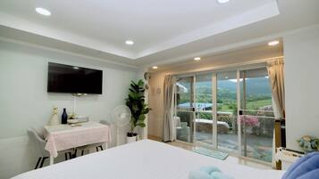 A bedroom with a bed, a TV, a table with a fan, and a view of a garden through a sliding door.