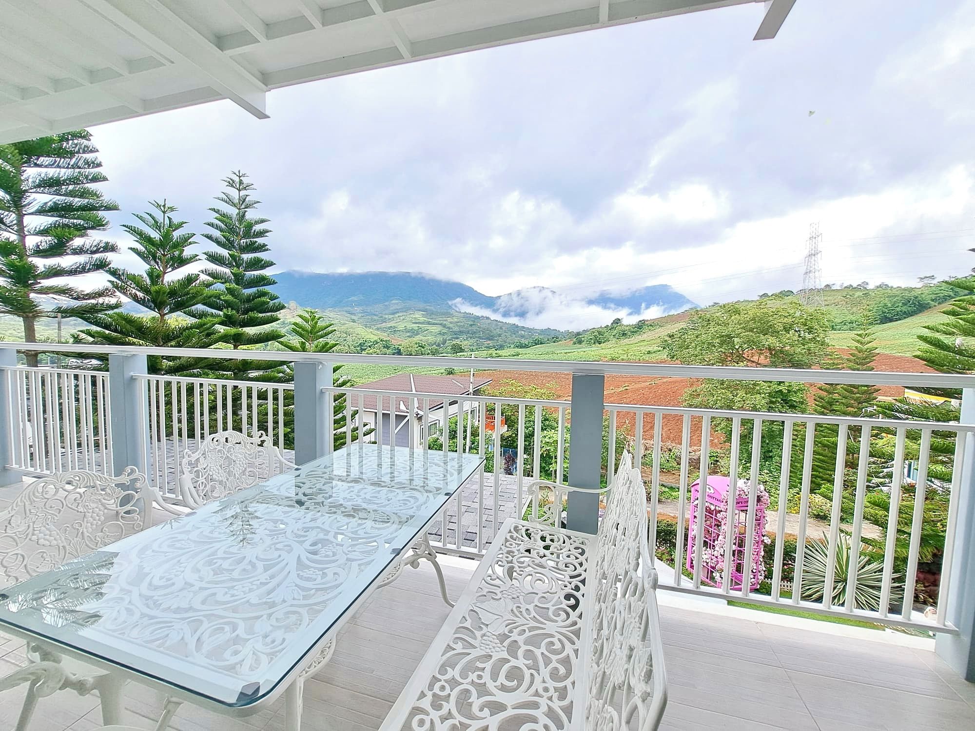 A balcony with a glass table and white ornate chairs, overlooking a green landscape with hills and a power line tower.