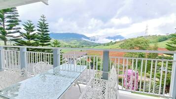 A balcony with a glass table and white ornate chairs, overlooking a green landscape with hills and a power line tower.