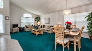 A spacious living room with a teal carpet, a dining area with wooden chairs, and a television on a stand. - Villa Near Disney 4 Bd 3 Bt With Private Pool Quiet Comminuty 4 Bedroom Home (Davenport)