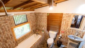 A bathroom with a bathtub, toilet, and sink, featuring stone walls and wooden beams. - Room-jungle Breeze Hideout (Ubud)