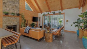 A spacious living room with a stone fireplace, wooden ceiling beams, and large windows.