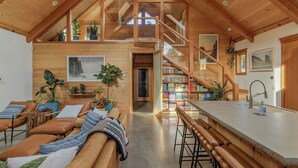 A spacious loft-style living area with a high ceiling, wooden beams, a mezzanine level, a kitchen with bar stools, and a staircase.