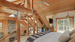 A spacious bedroom with a large bed, wooden ceiling beams, a stone fireplace, and large windows offering a view of the outdoors.
