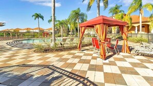 Un área junto a la piscina con suelo a cuadros, un espacio de descanso cubierto con sillas y palmeras.