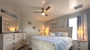 Un dormitorio con cama, mesitas de noche, cómoda, ventilador de techo, decoración en la pared y una ventana con cortinas.