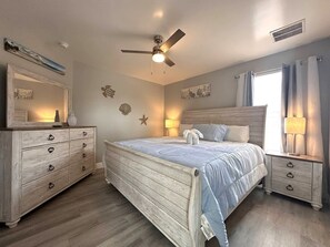 Ein Schlafzimmer mit Bett, Nachttischen, einem Schminktisch, einem Deckenventilator, Wanddekoration und einem Fenster mit Vorhängen.