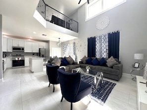 A modern living room with a grey sofa, two armchairs, and a coffee table. There is a kitchen area with white cabinets and a stainless steel refrigerator. A balcony with a black railing is visible above the living room. - Luxury Villa Near Disney 6 Bd Home With Private Pool And Resort 6 Bedroom Home (Davenport)