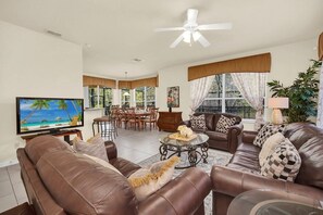 A living room with a television, leather sofas, a dining area with a table and chairs, and large windows with curtains. - Near Disney Amazing 5 Bd Home - Private Pool 5 Bedroom Home (Davenport)
