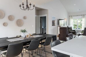 A modern dining area with a dark wooden table, black chairs, and a chandelier. There are decorative wall pieces and a window with blinds. - Laurel Hill Farmhouse | Private Hot Tub, Fire Pit, One-Level Modern Farmhouse Near Asheville (Fletcher)