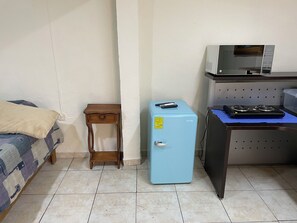 Una habitación pequeña con una cama, una mesita de noche de madera, un refrigerador azul, un escritorio con una computadora y un monitor, y un calentador fijado a la pared.