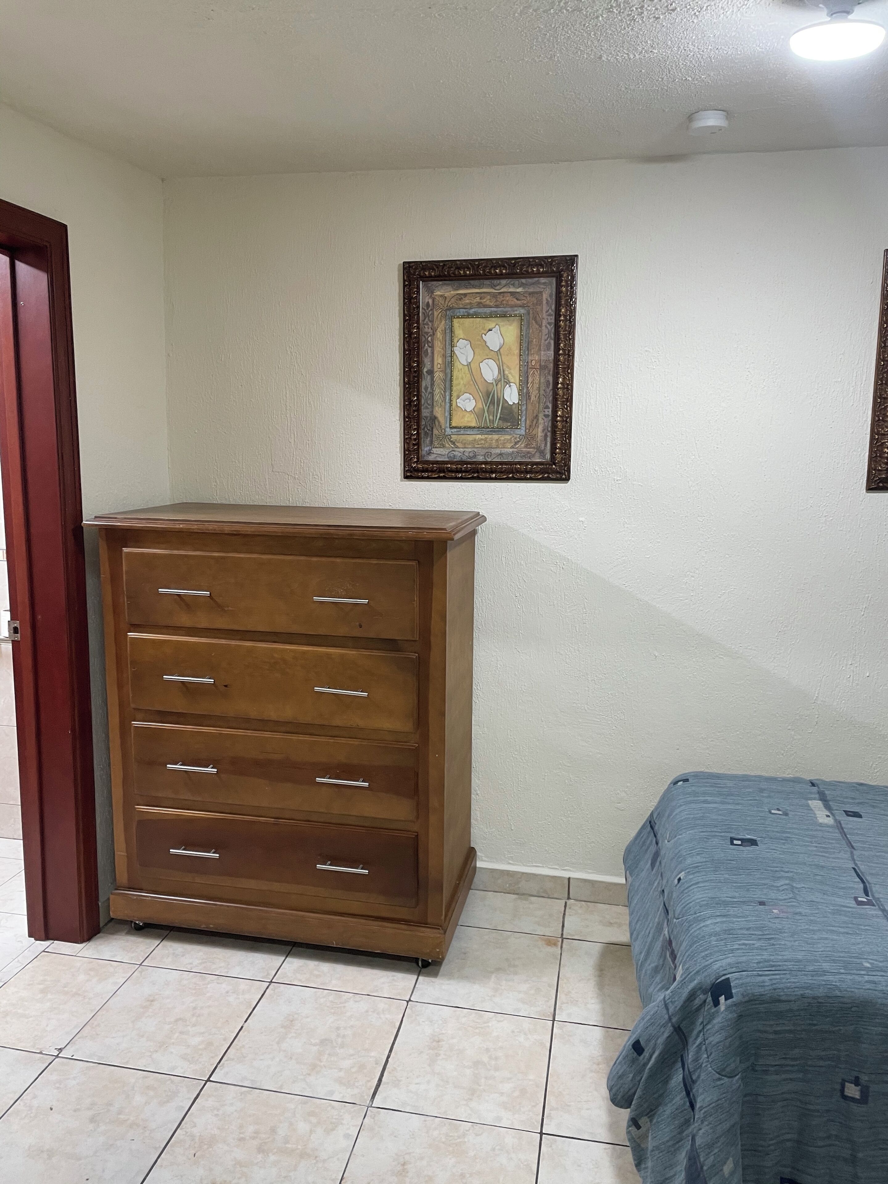 A room with a wooden dresser, a bed with a grey bedspread, and a framed picture on the wall.