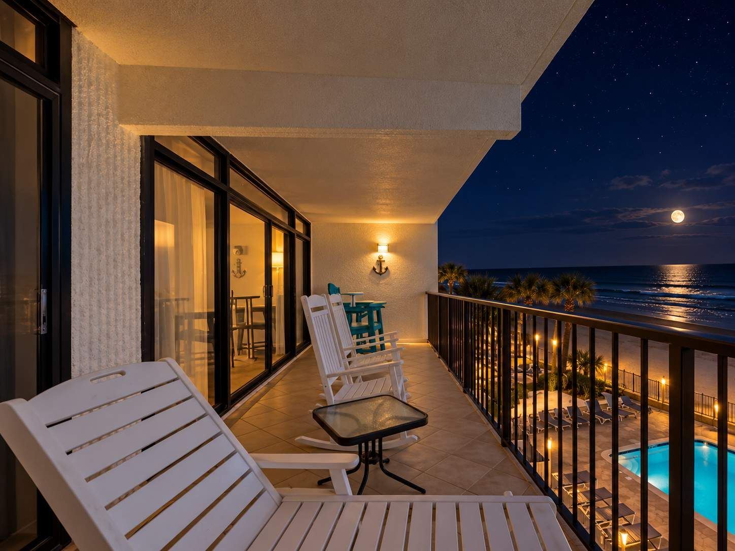 A balcony with white chairs and a table, overlooking a pool and the ocean at night.