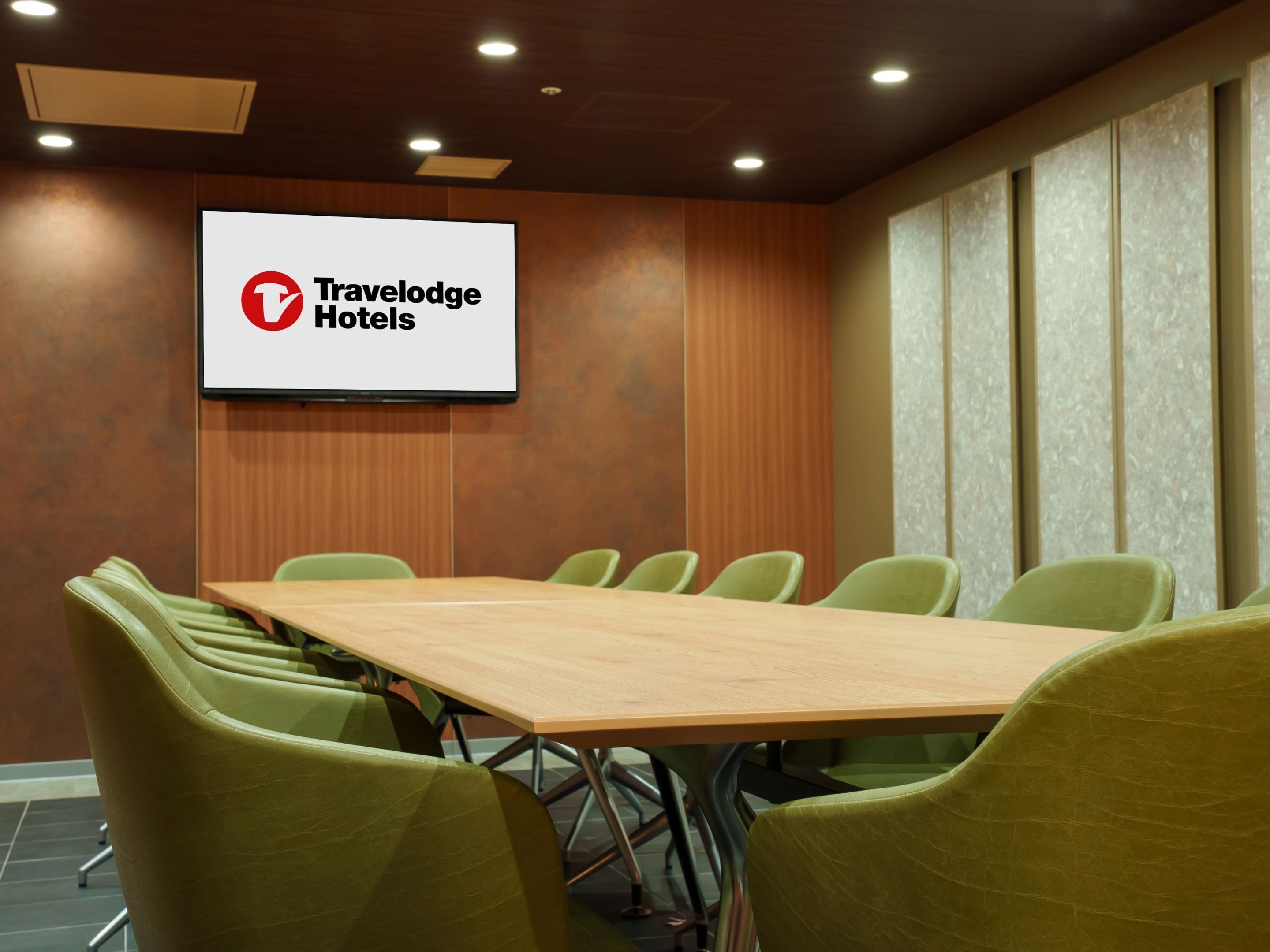 A conference room with a long table and six chairs, a screen displaying 'Travelodge Hotels' logo, and wooden panel walls.