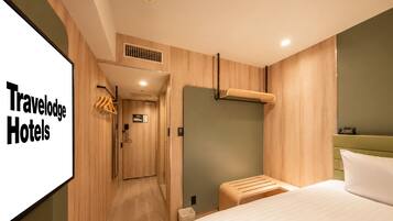 A modern hotel room with a wooden floor, a bed with white bedding, a small bedside table, a wall-mounted heater, and a wardrobe with a mirror.