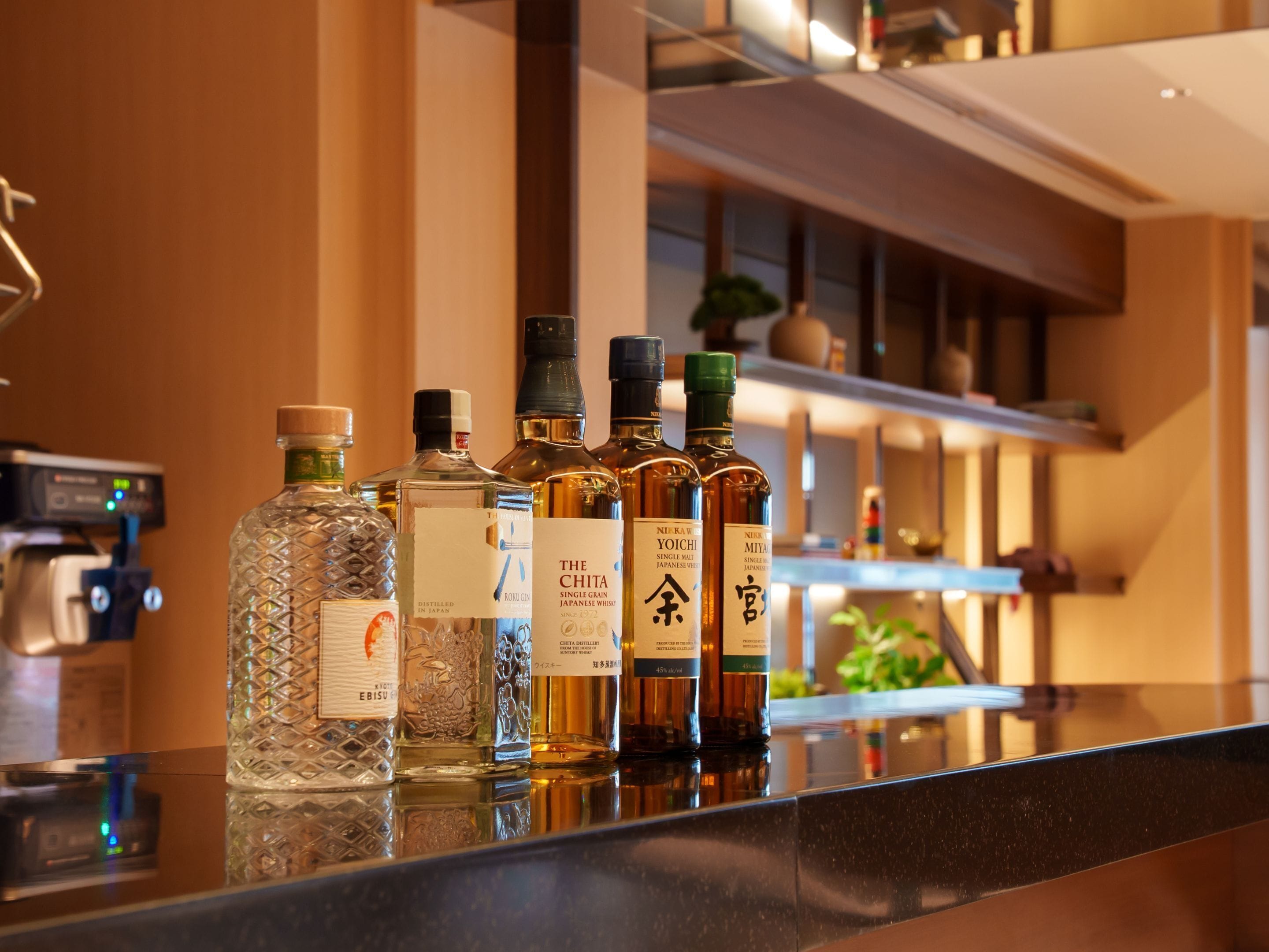 Uma área de bar com várias garrafas de bebidas alcoólicas, incluindo uísque e saquê, sobre uma superfície refletora.