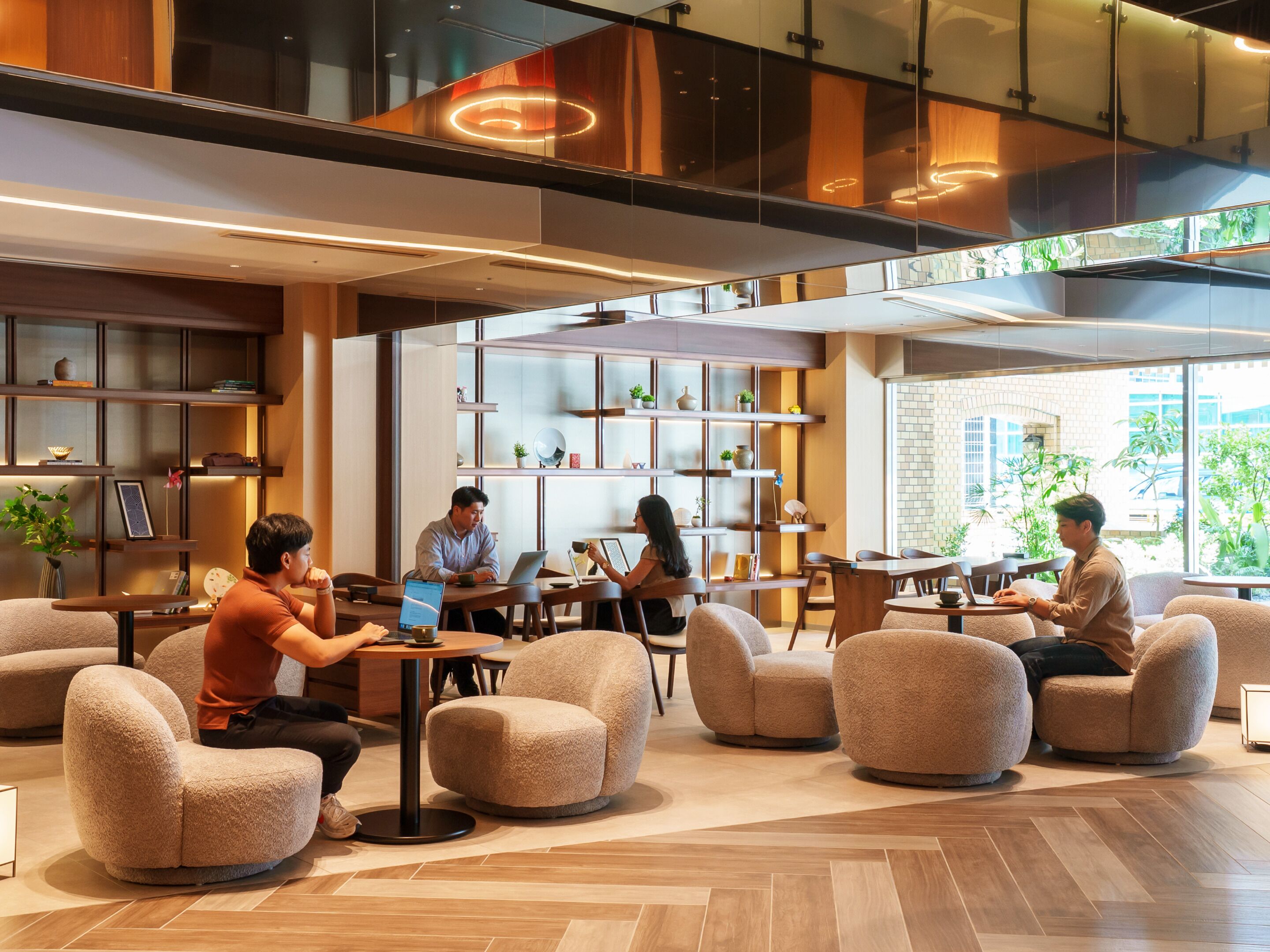 A modern indoor space with round tables and comfortable seating, a large bookshelf, and a view of greenery outside.