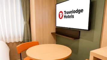 A hotel room with a bed, a round table, two orange chairs, a television mounted on the wall, and a wooden shelf below the TV.