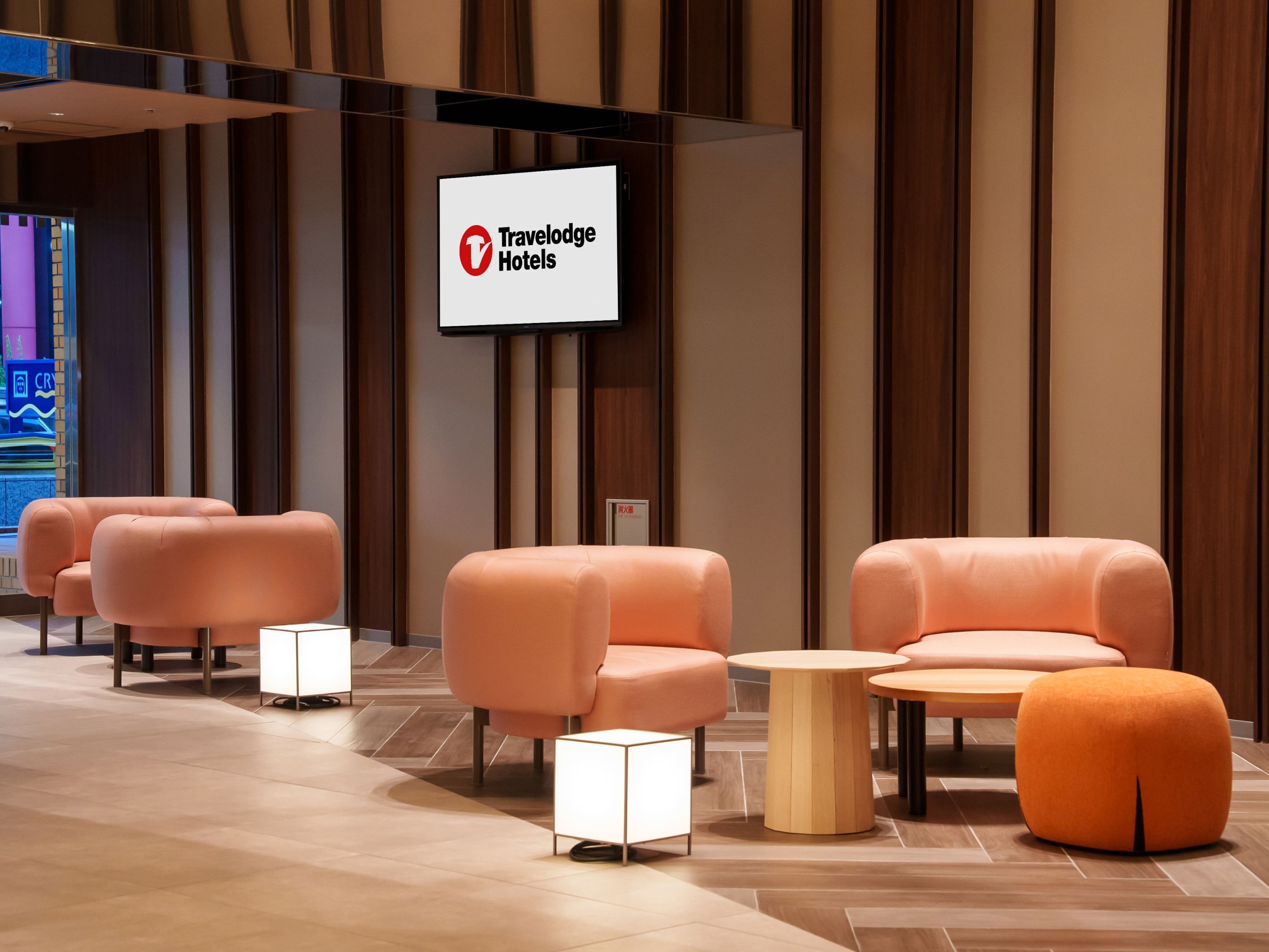A modern hotel lobby with plush seating, a central round table, and a large sign for Travelodge Hotels.