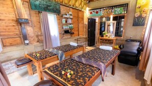 Una habitación con paredes y suelos de madera, dos camillas de masaje con fundas estampadas, una pequeña mesa de madera con un espejo y una silla de masaje negra.