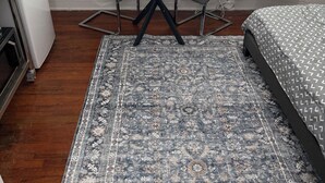 Une pièce de taille modeste, avec une table ronde et deux chaises, un lit recouvert d’une literie à motifs, une grande moquette, une fenêtre avec des stores et un appareil de climatisation.