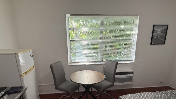 Une petite pièce meublée, comprenant un lit, une table, deux chaises, un réfrigérateur et une fenêtre avec des stores.