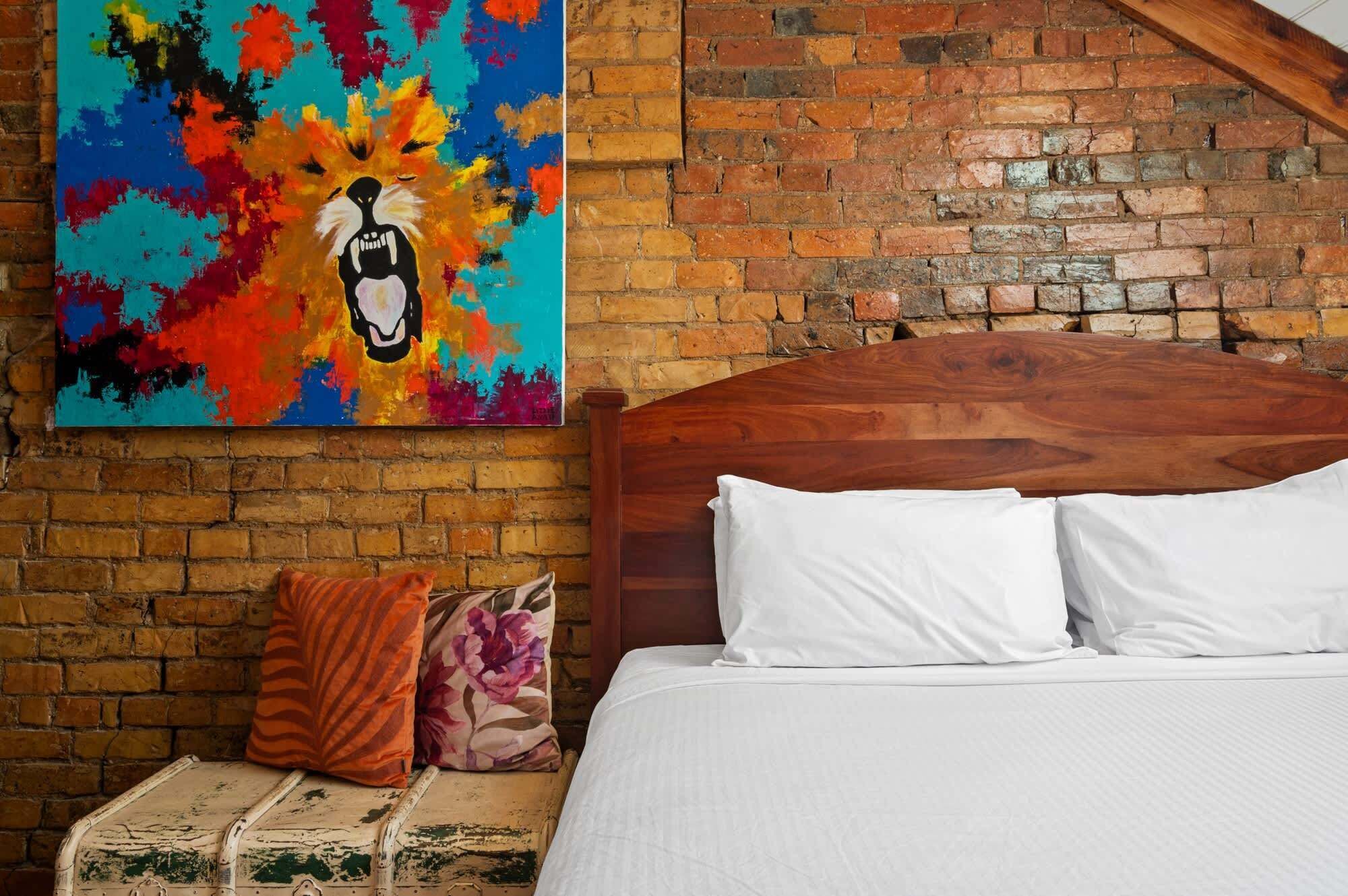 A bedroom with a wooden headboard, white bedding, a colorful abstract painting, and a rustic brick wall.