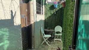Um corredor exterior estreito com chão em azulejos, uma cadeira branca e uma mesa pequena.