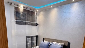 Quarto com cama, janela com cortinas, luminária no teto e tapete estampado.