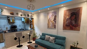 Uma sala de estar moderna com um sofá azul-turquesa, uma mesa de centro e um tapete estampado. Uma cozinha com armários e bancada é visível ao fundo.