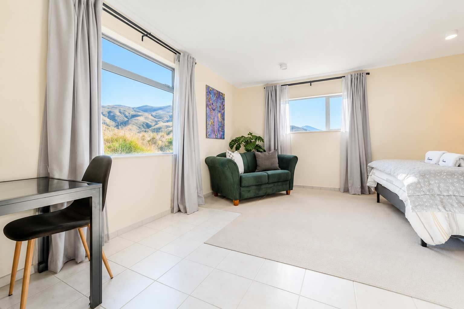 Una habitación moderna con cama, sofá verde, escritorio con silla y ventanas grandes con cortinas.