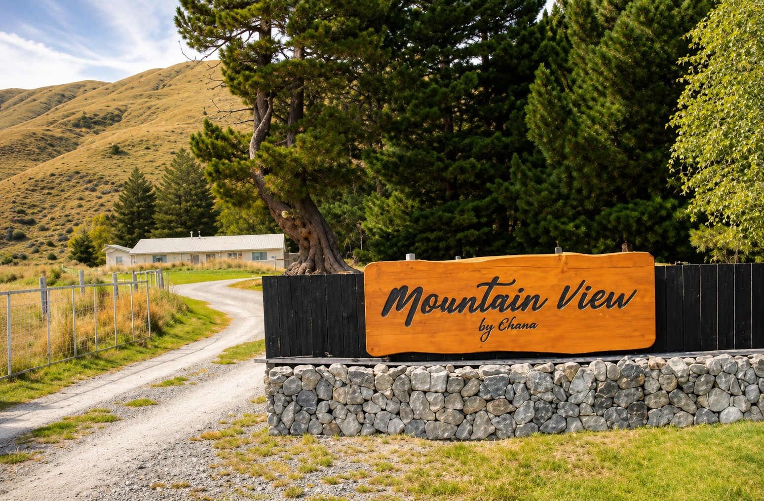 A wooden sign with 'Mountain View by Chana' written on it, a gravel path, a fenced area, and a building in the background.