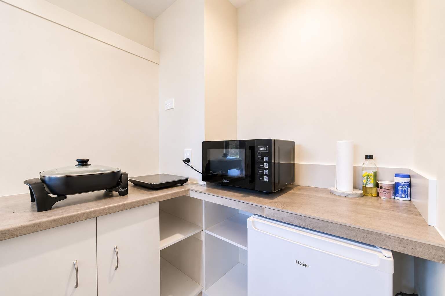 Una kitchenette equipada con microondas, cocedor de arroz, un refrigerador pequeño y una encimera con una botella de aceite y un recipiente con harina.