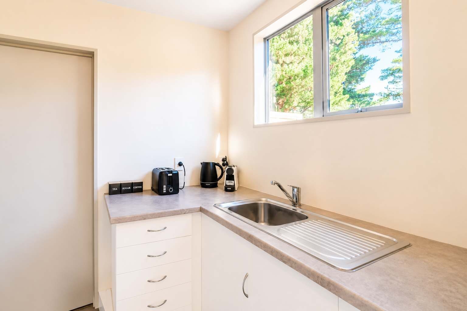 A compact kitchenette with a sink, a coffee maker, a toaster, and a window with a view of trees.