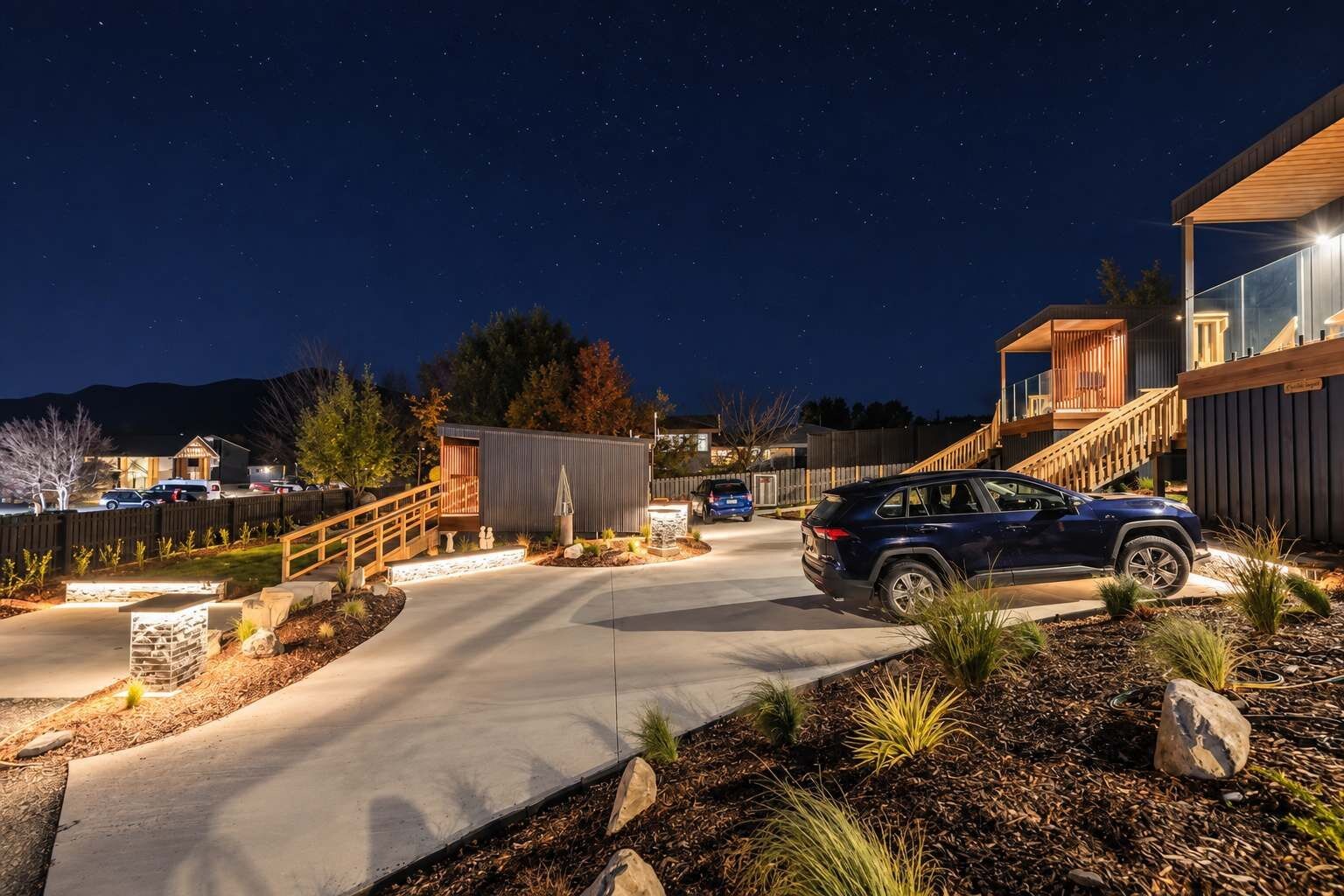 A modern residential property at night with a driveway, a blue SUV parked, and a wooden staircase leading to a second-story deck.