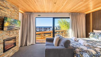 Une chambre d’hôtel avec une cheminée, un canapé, un lit avec une literie à motifs floraux et une vue sur une terrasse en bois et les bâtiments environnants.