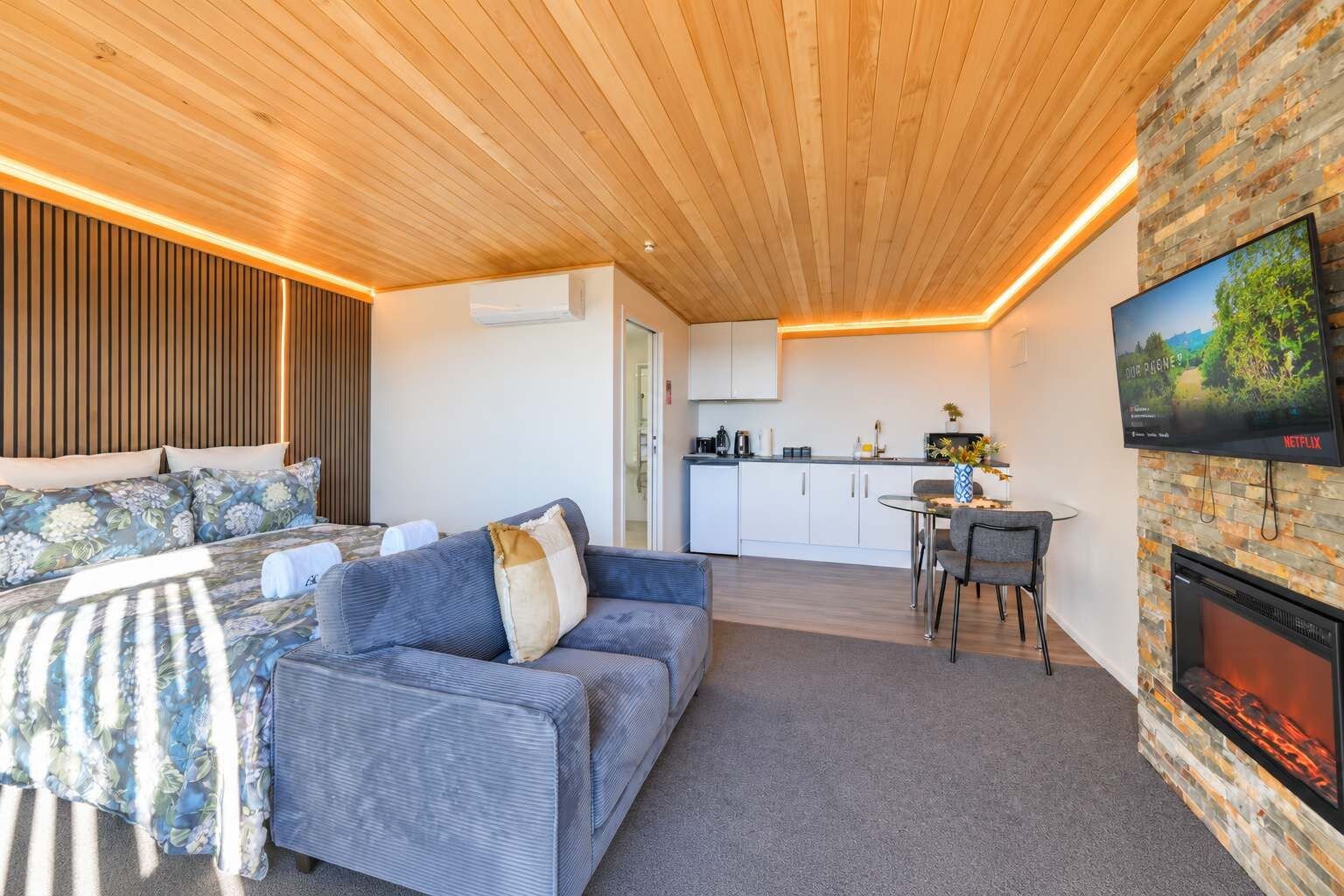 A modern living room with a sofa, a flat-screen TV mounted on a stone wall, and a fireplace.
