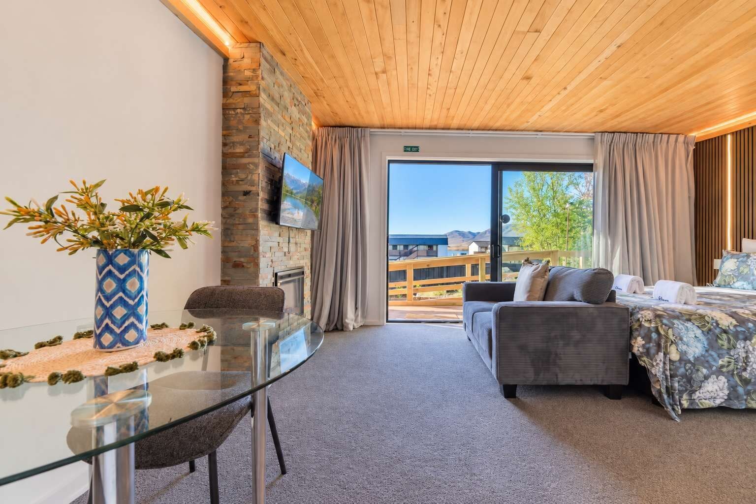A hotel room with a glass table, a sofa, a bed with floral bedding, a fireplace, and a view of the outdoors.