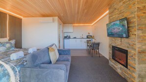 Une pièce moderne avec un plafond en bois, une cheminée, un téléviseur fixé au mur et, au fond, un coin cuisine.