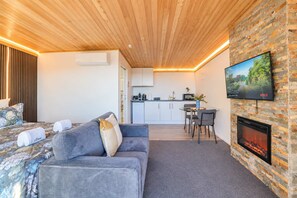 A modern room with a sofa, a flat-screen TV mounted on a brick wall, a fireplace, and a kitchen area with cabinets and a countertop. - Skyline Haven | Lake Tekapo + Lake Views (Lake Tekapo)