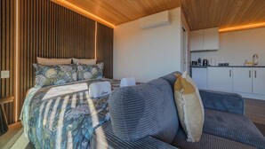 Une chambre moderne avec un plafond en bois, un lit recouvert d’une literie à motifs floraux, un canapé gris et un coin cuisine.